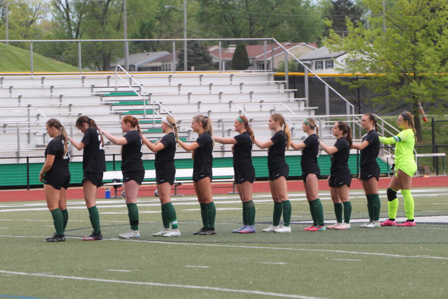 Starters for the girls varsity soccer team stand together with their hands on each others shoulders during the national anthem before their senior night game.